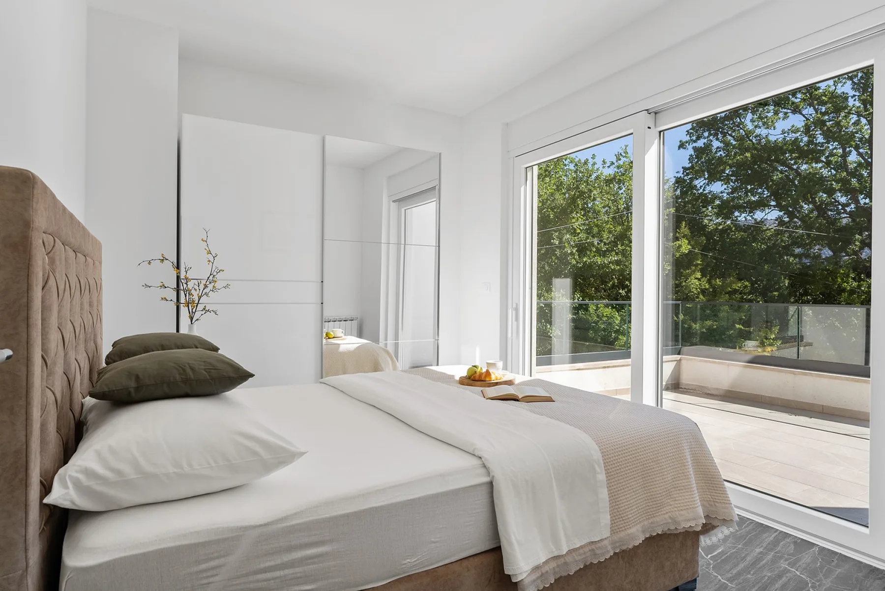 A bright bedroom with a bed beside large windows overlooking leafy trees.