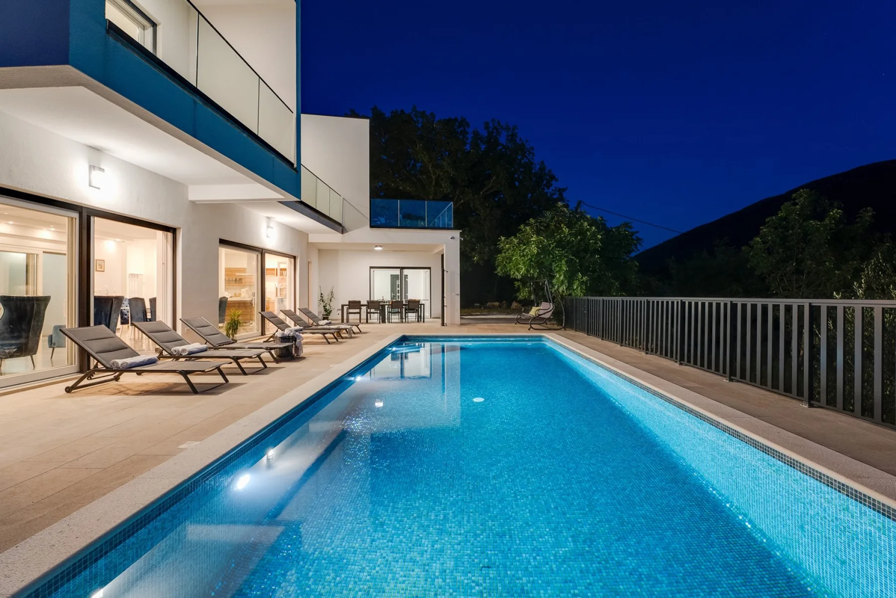 An outdoor swimming pool beside a modern building, with lounge seating on the patio.