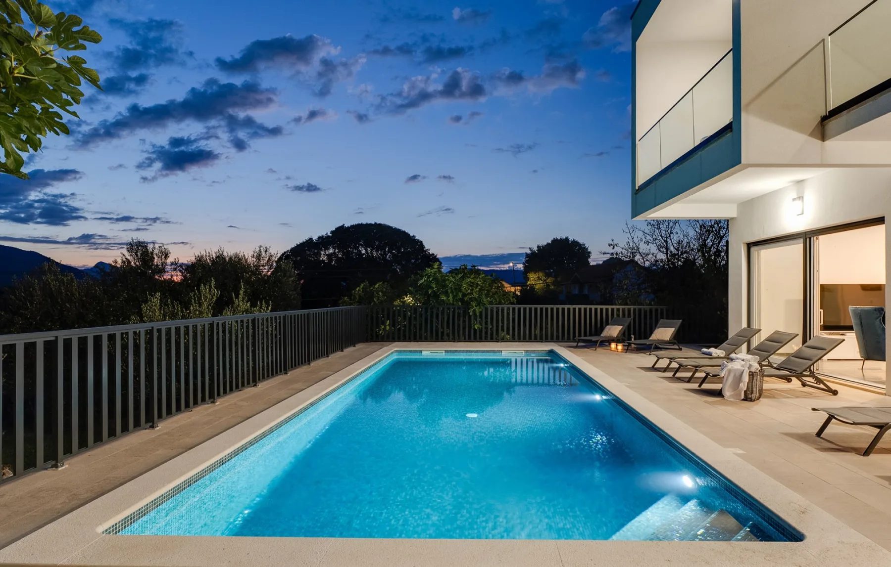 An outdoor swimming pool beside a modern building at dusk, with lounge seating along the deck.