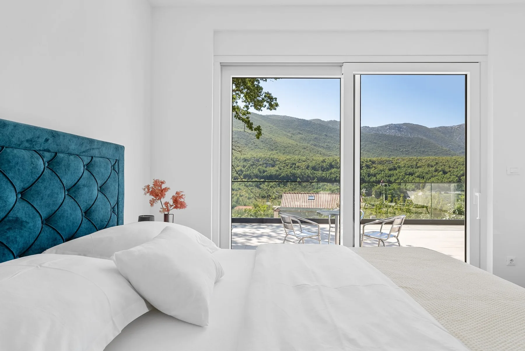 A bright bedroom opens onto a terrace with seating and views of green hills.