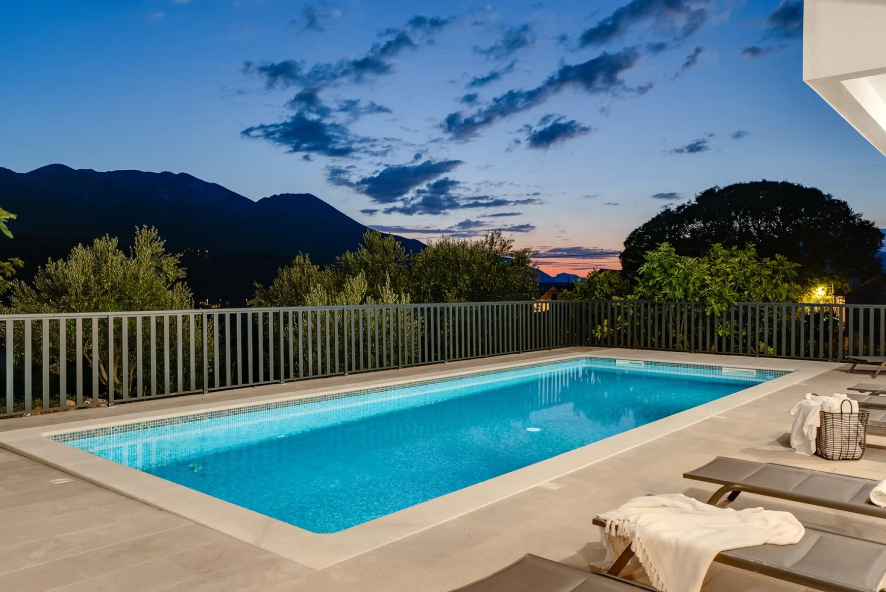 An outdoor swimming pool and lounge seating overlook a mountain landscape at sunset.