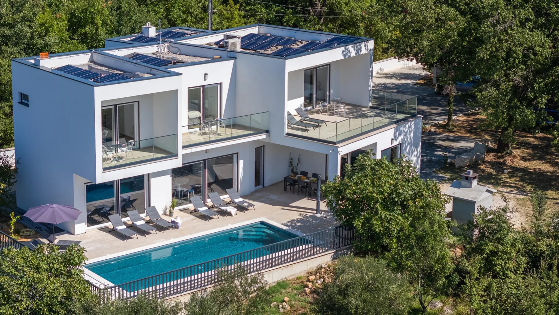 A modern white house overlooks a swimming pool, with trees surrounding the property.