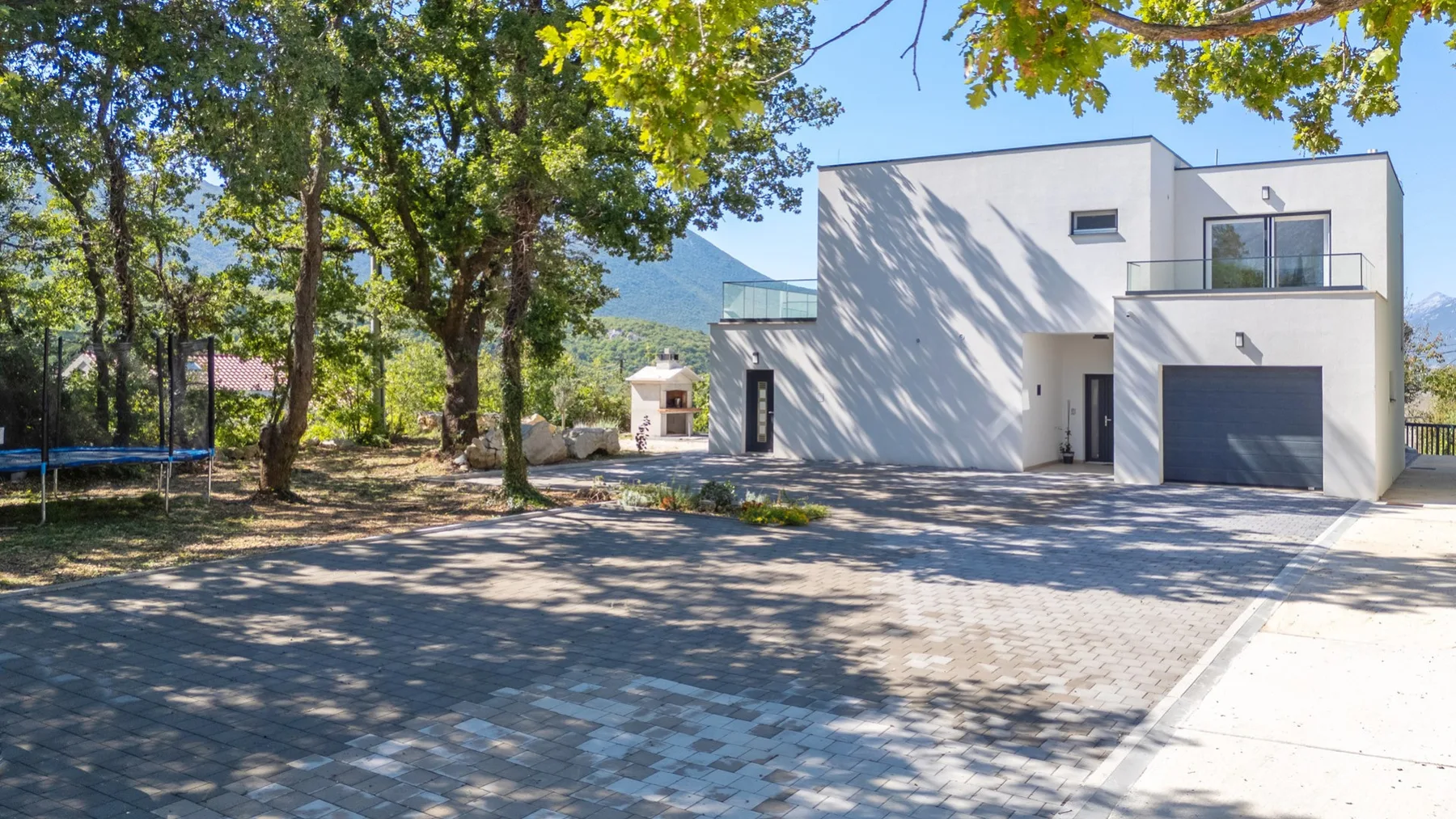 A modern white house stands beside a paved driveway beneath leafy trees.