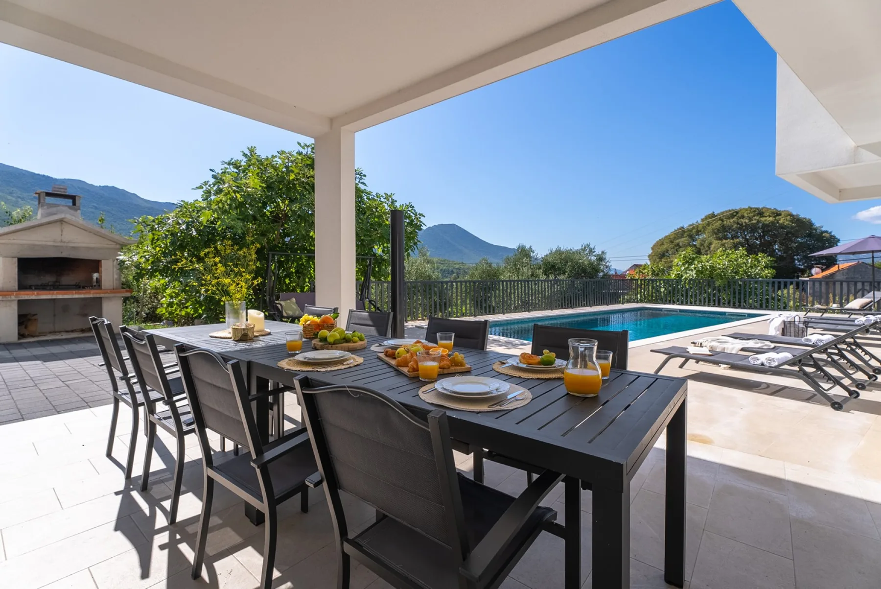 An outdoor dining area overlooks a swimming pool and surrounding hills.