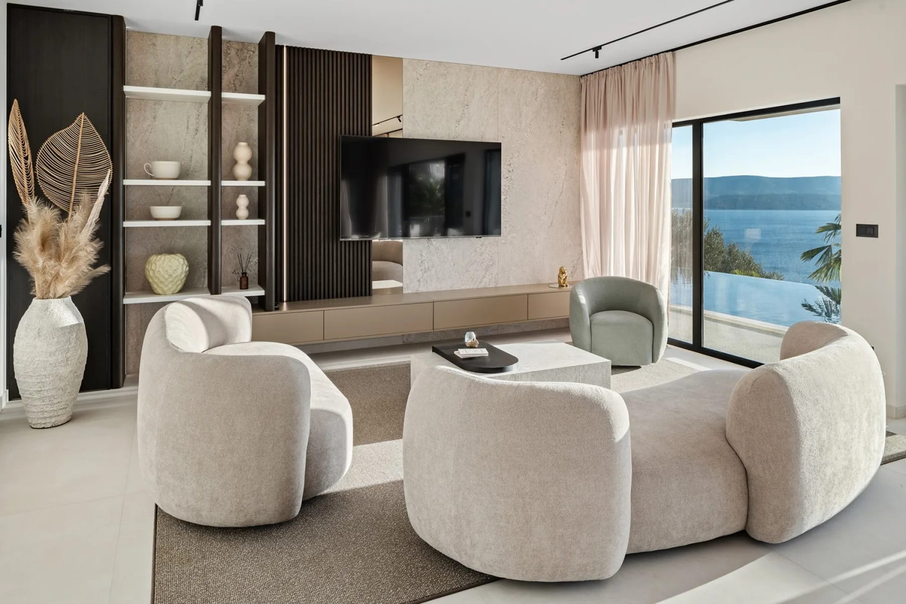 A modern living room with curved seating, a television, and a large window overlooking the water.