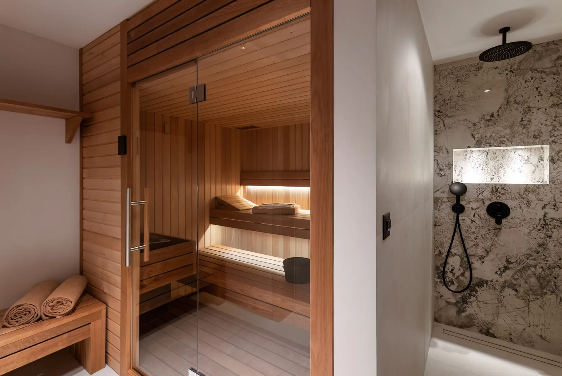 A wooden sauna with tiered seating stands beside a tiled shower.