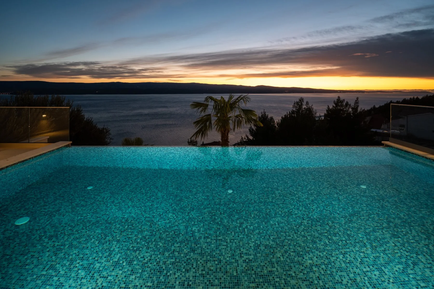 An infinity pool overlooks a lake and distant hills at sunset.