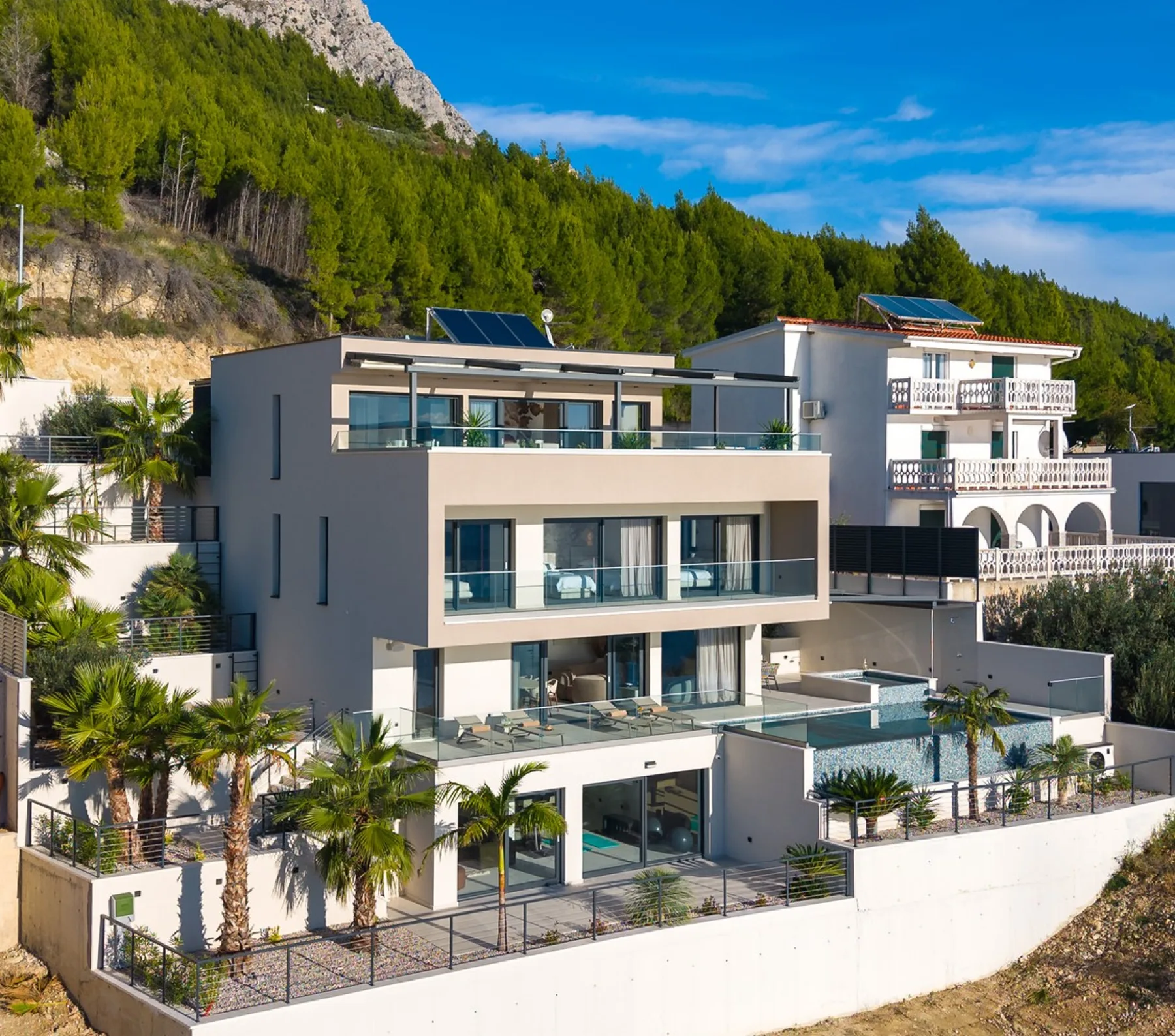 A modern house with balconies and a swimming pool sits below a wooded hillside.