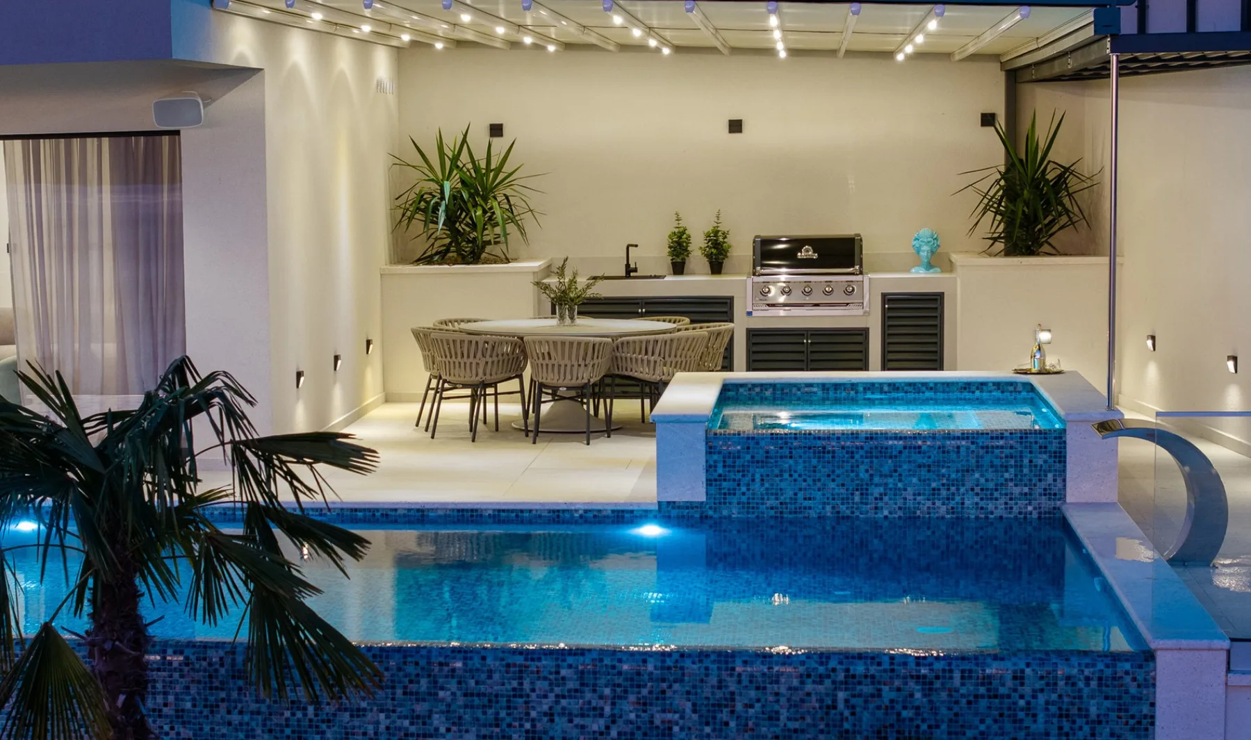 A swimming pool sits beside an outdoor dining area beneath string lights.