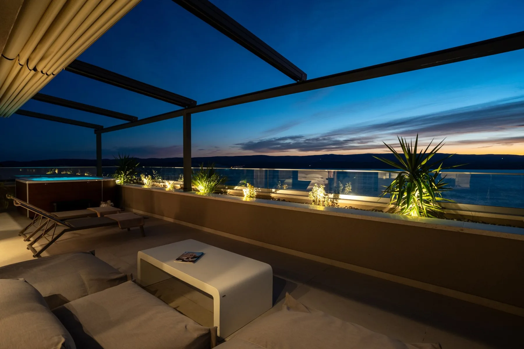 A rooftop terrace overlooks the water at dusk, with lounge seating beneath a pergola.