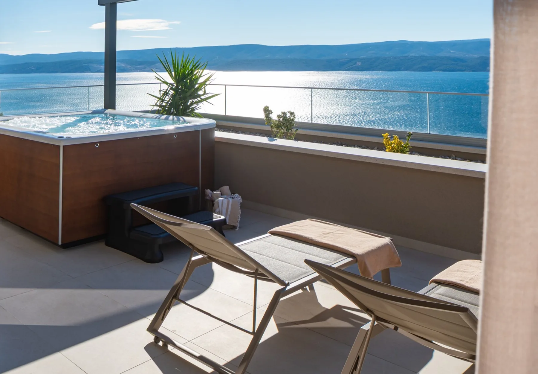 A terrace with lounge seating and a hot tub overlooks the water and distant hills.