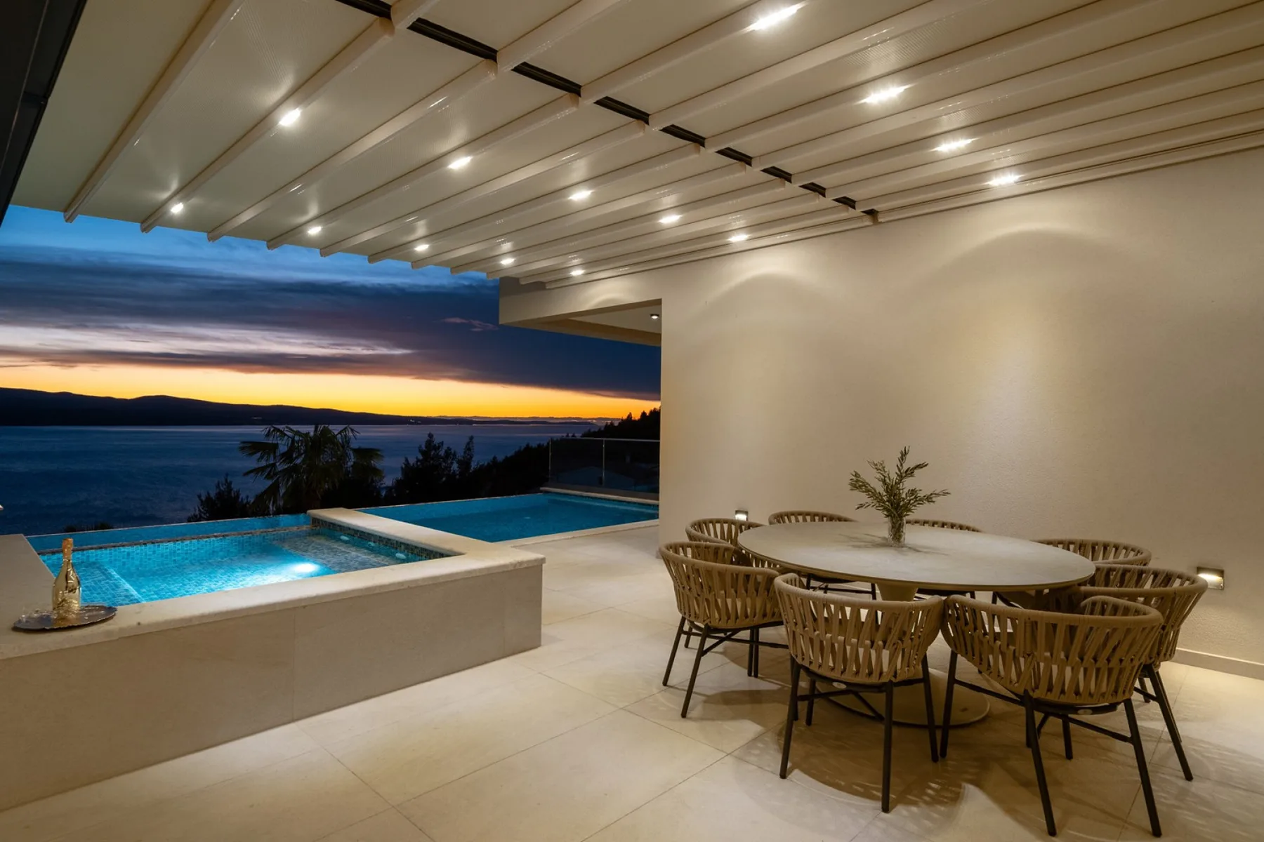 A covered outdoor dining area overlooks a pool and the sea at sunset.