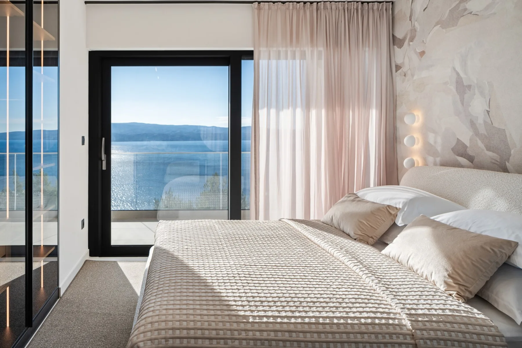 A bright bedroom with a neatly made bed beside a large window overlooking the water.