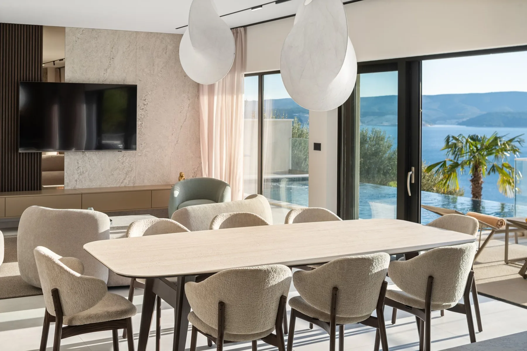A bright dining area with a long table and upholstered seating beside floor-to-ceiling windows.