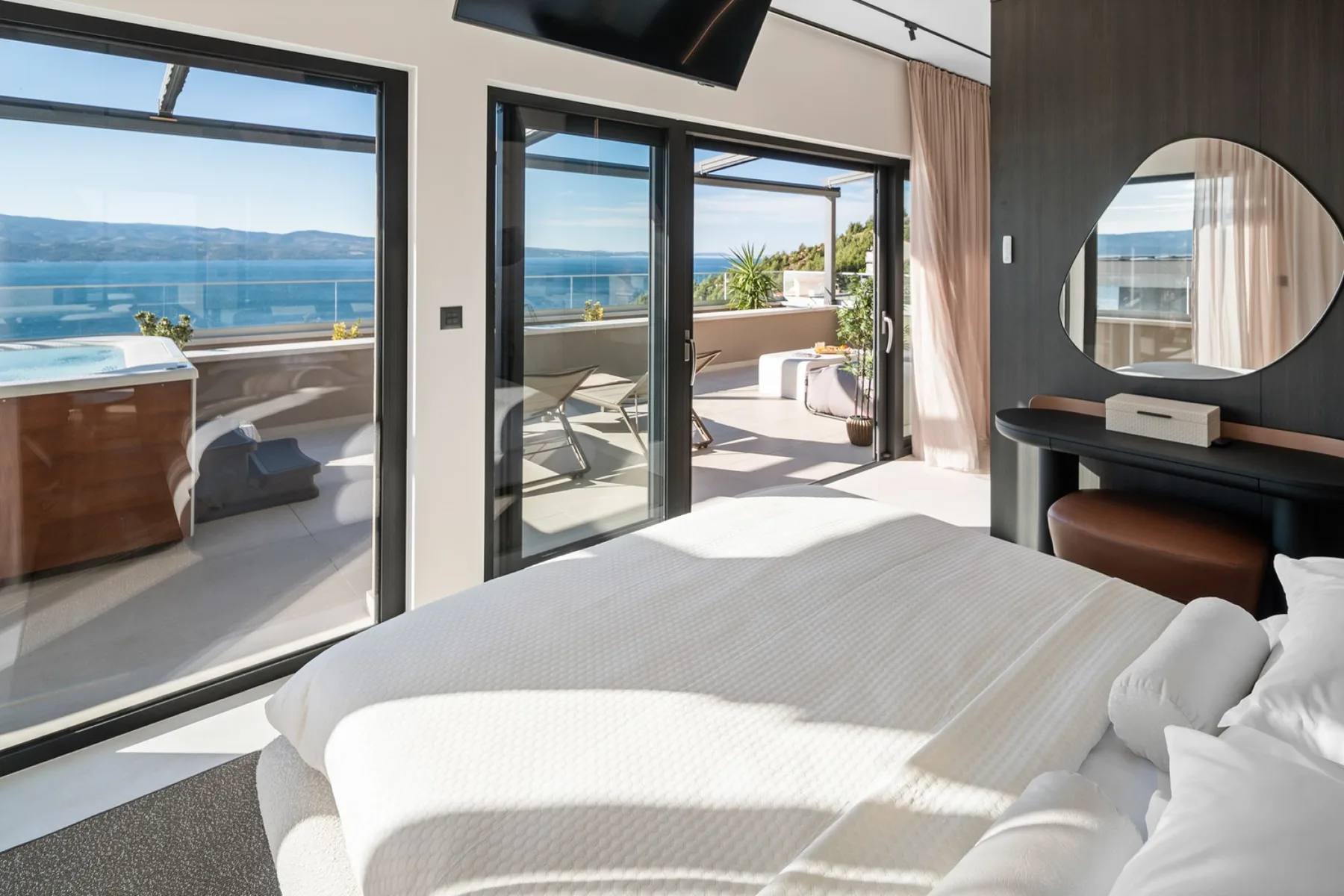 A bright bedroom with a bed, a round mirror, and large glass doors overlooking the water.