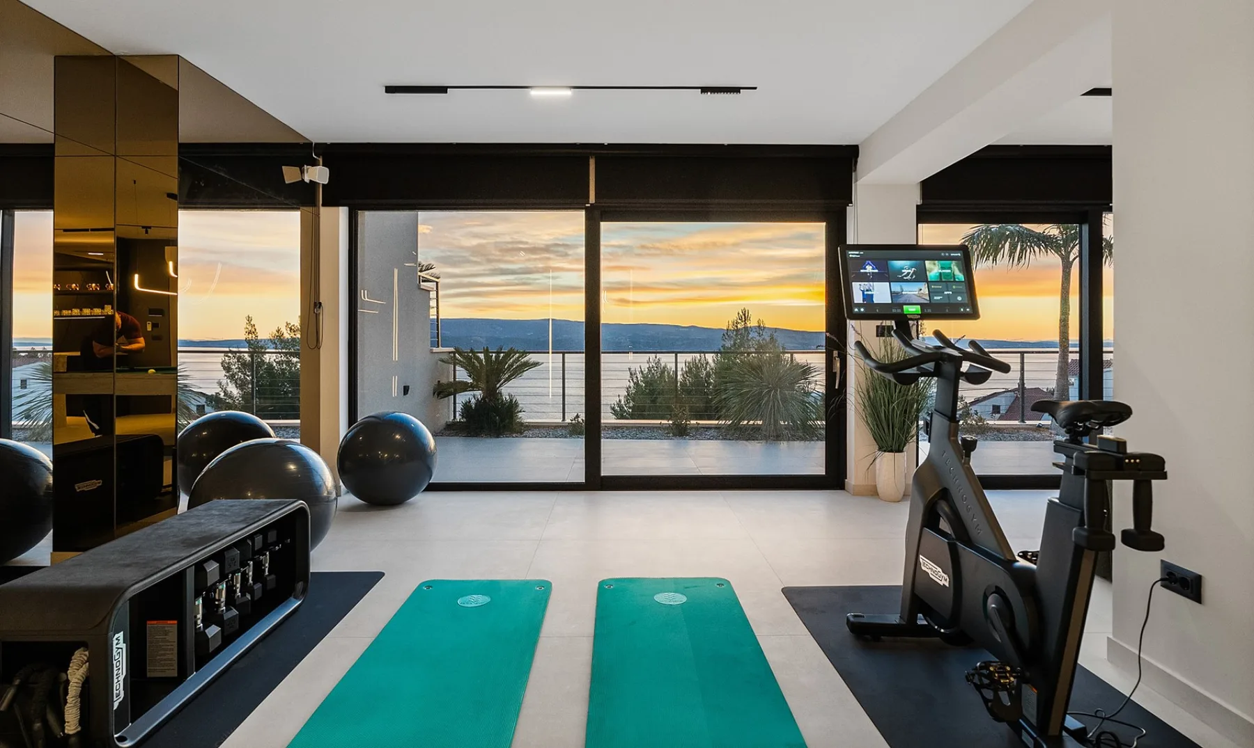 A bright fitness room with exercise equipment and a sunset view through the windows.