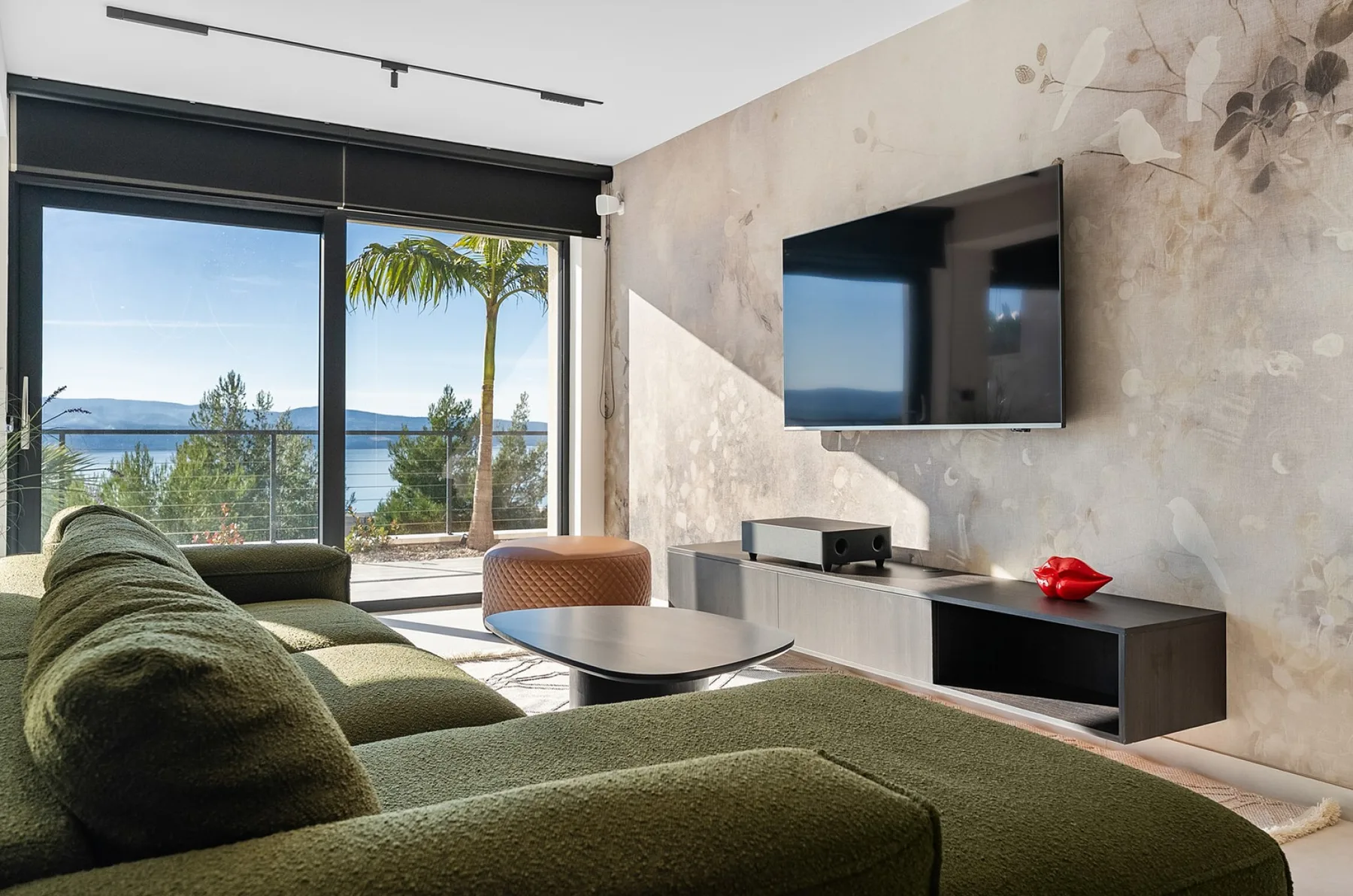 A living room with green seating, a wall-mounted television, and sliding glass doors overlooking the water.