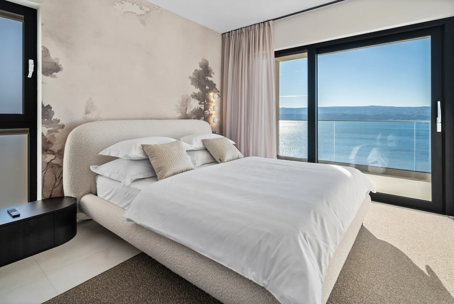 A bedroom with a neatly made bed beside large windows overlooking the water.