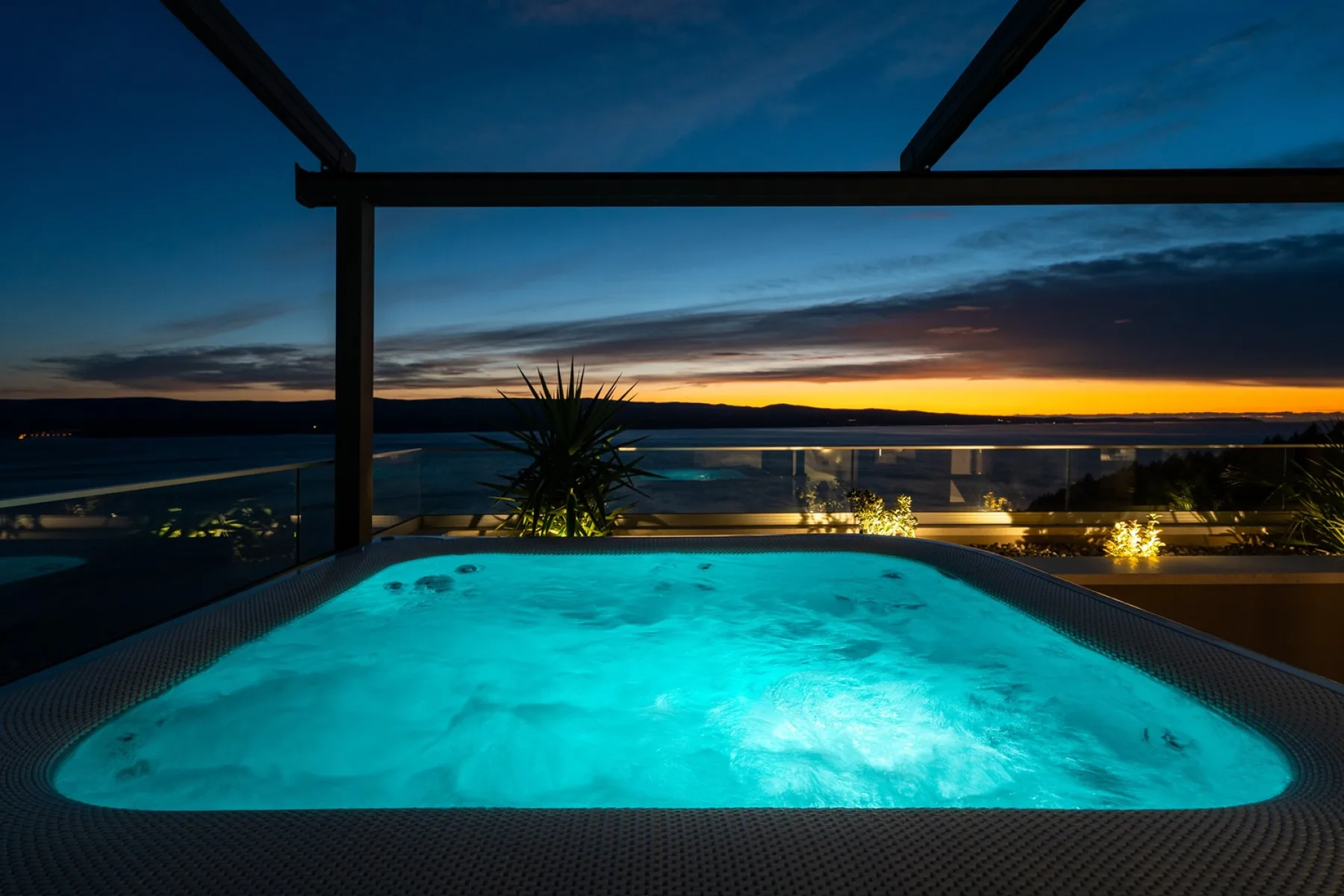 An illuminated outdoor hot tub overlooks a coastal sunset.