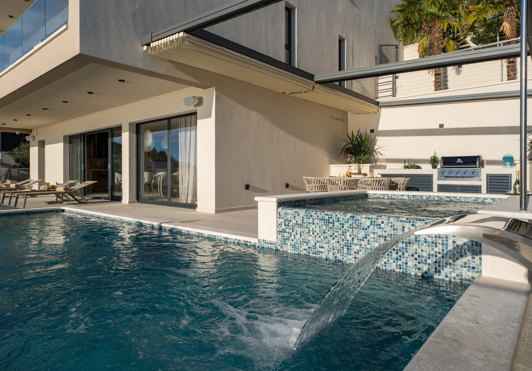 A swimming pool beside a modern house, with a tiled water feature.