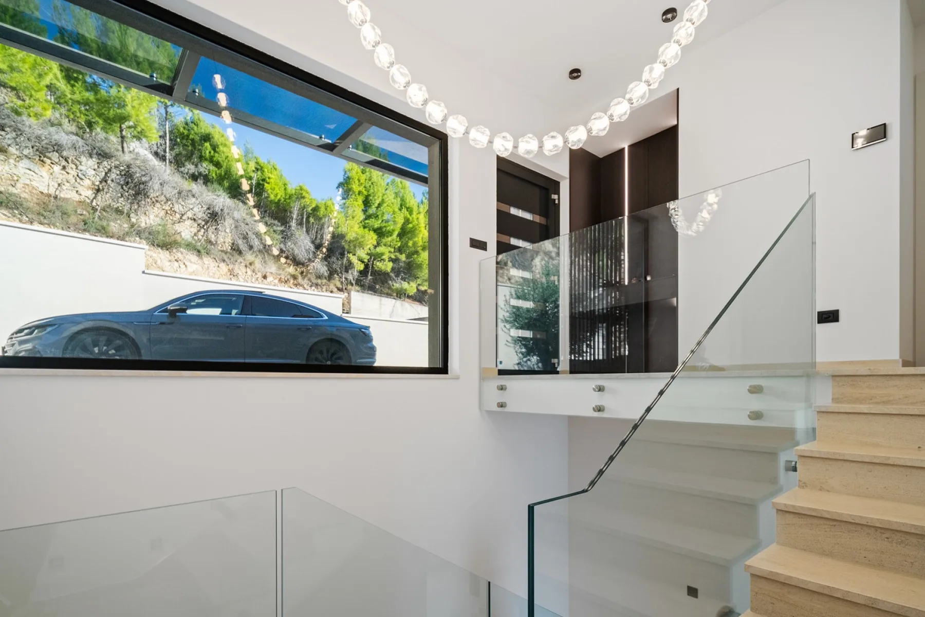 A bright modern interior features a staircase with a glass railing beside a large window.
