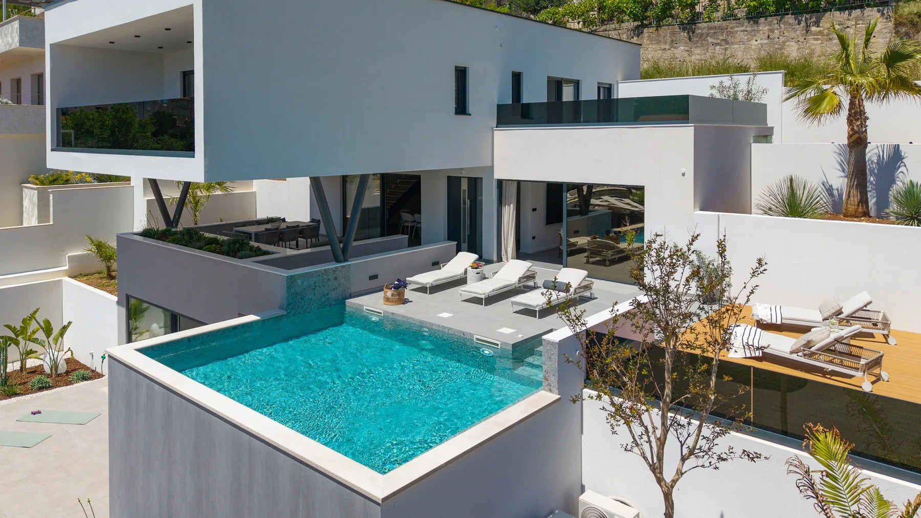 A modern white house overlooks a turquoise swimming pool and outdoor lounge area.
