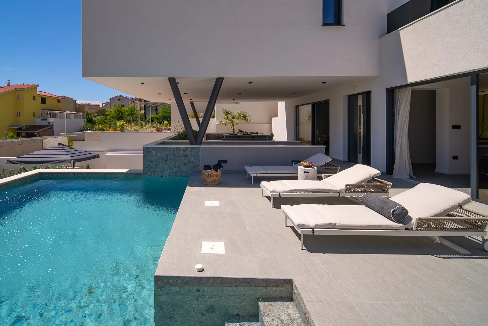 A modern house opens onto a swimming pool and a sunlit terrace with lounge seating.