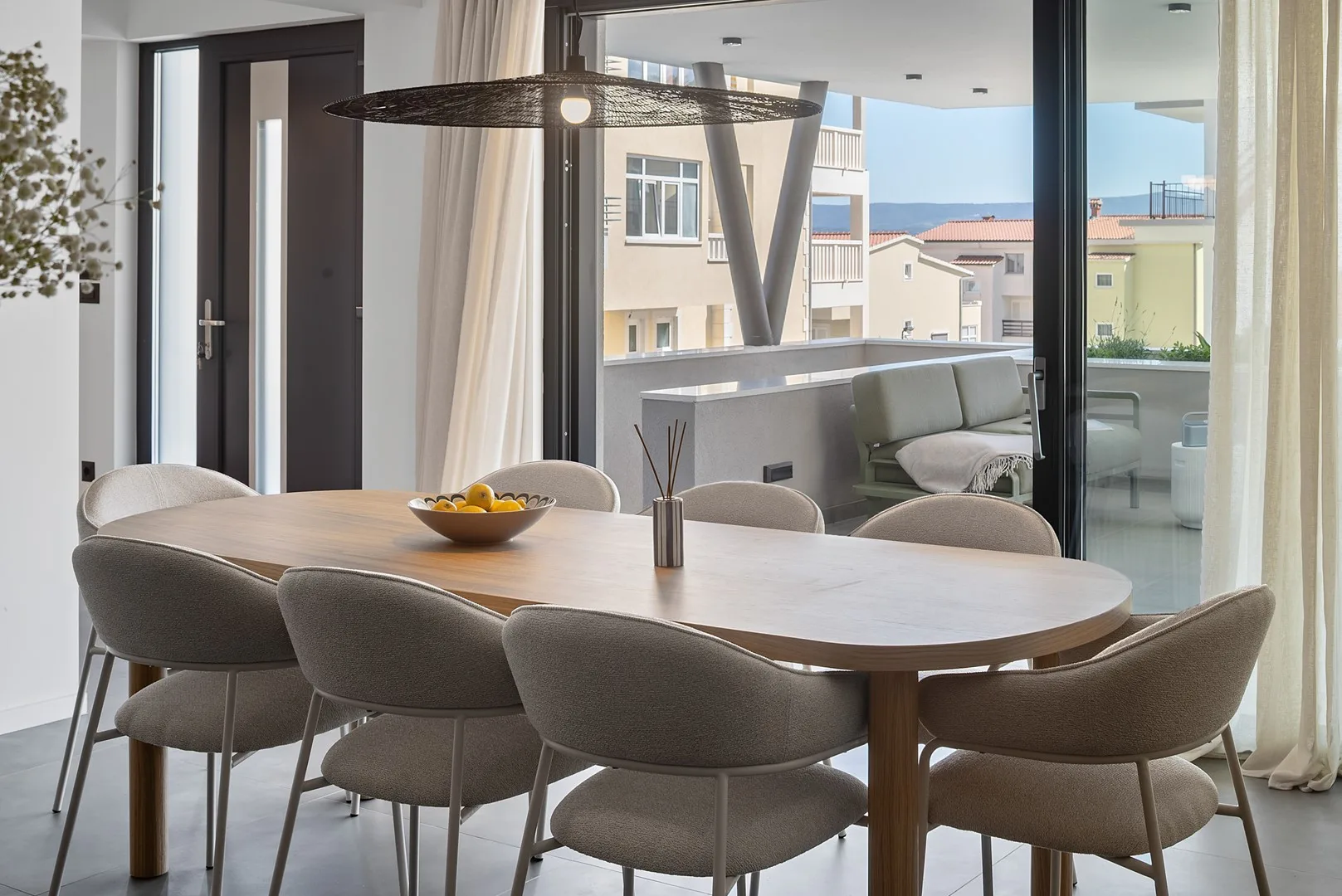 A dining area with a long table and upholstered seating beside large windows.