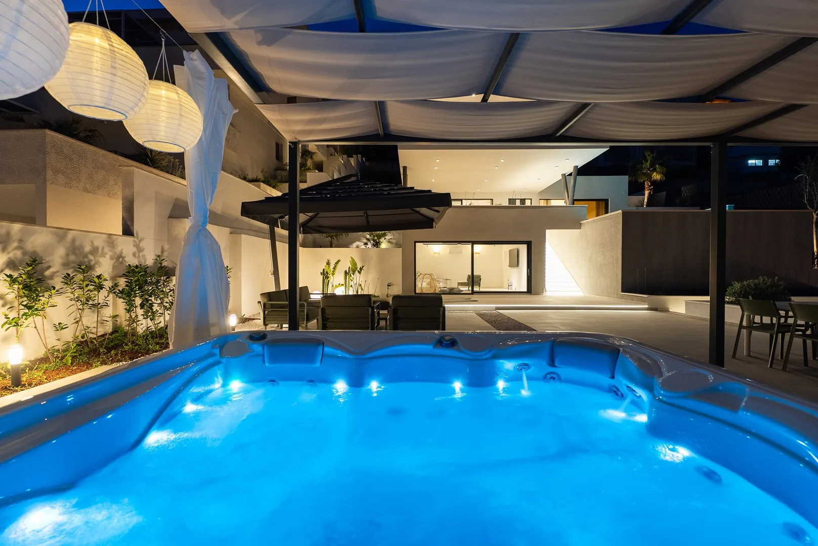 A glowing outdoor pool sits beneath a shaded canopy beside a modern building.