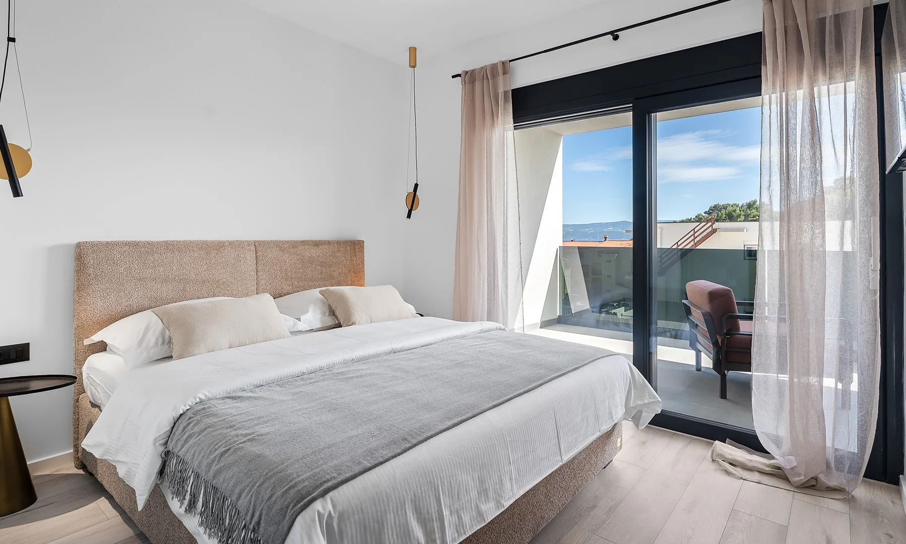 A bright bedroom with a neatly made bed beside glass doors opening onto a balcony.
