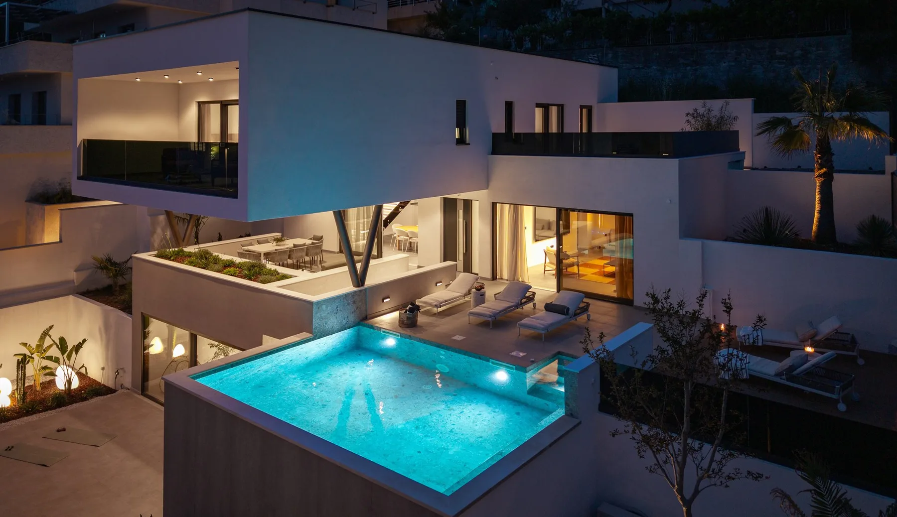 A modern house at night overlooks a lit swimming pool and outdoor seating.