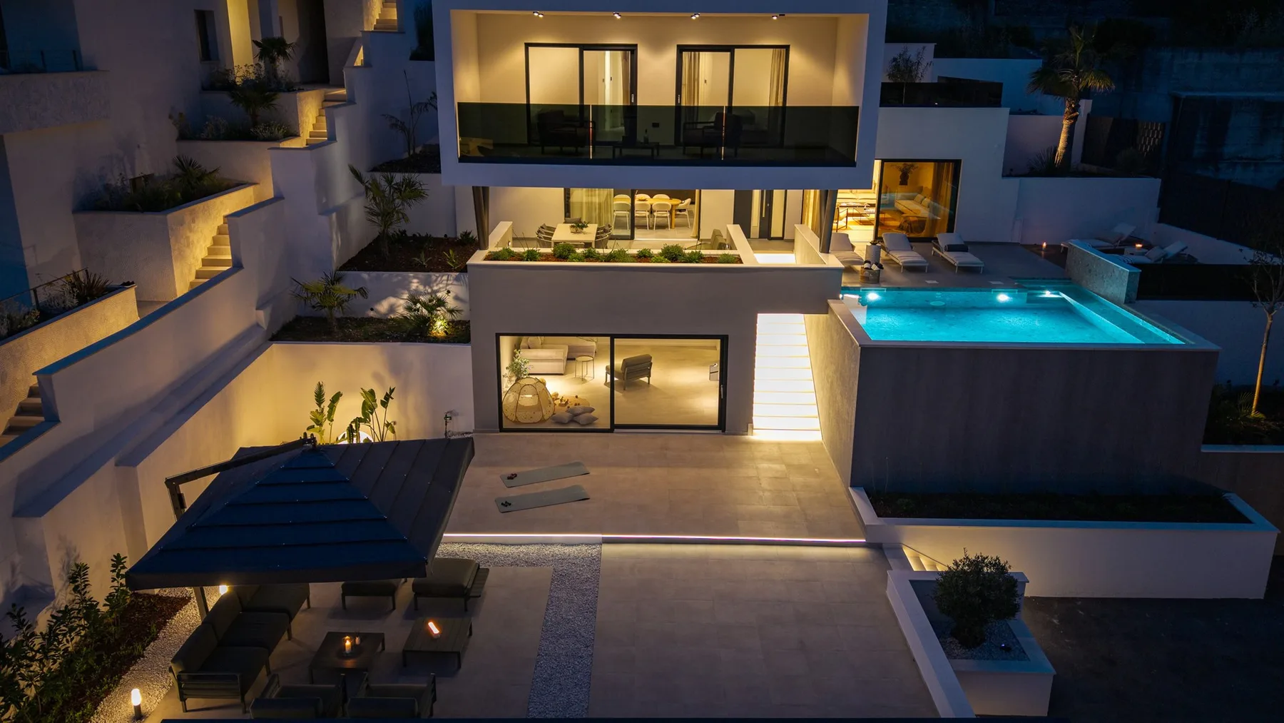 A modern multi-level building at night overlooks a brightly lit swimming pool and courtyard.