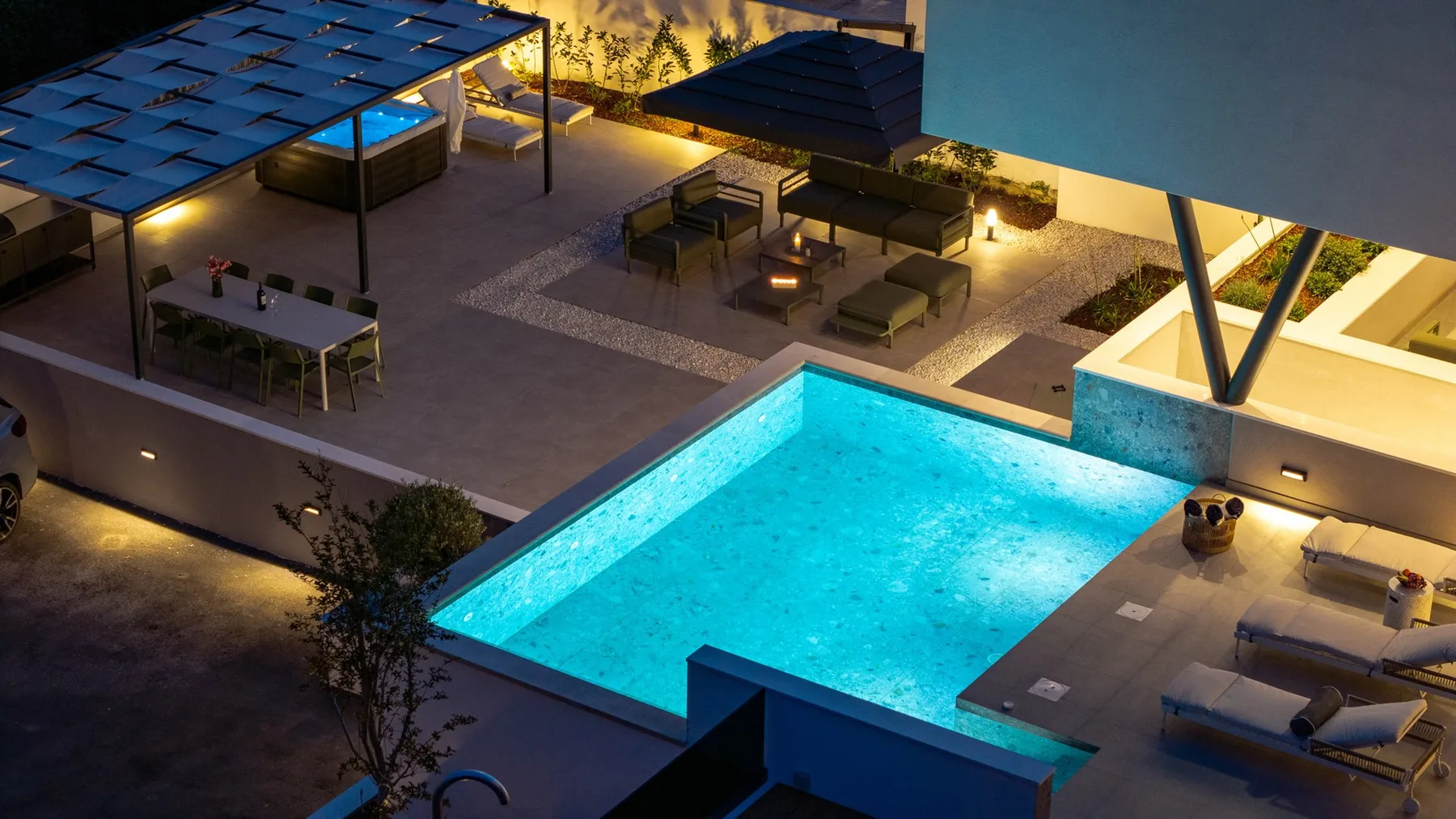 An illuminated swimming pool sits beside a furnished rooftop terrace at dusk.