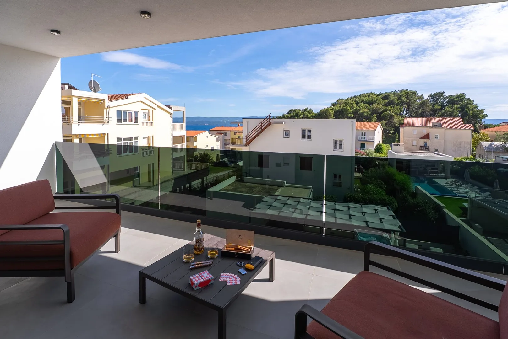 A balcony seating area overlooks nearby buildings and a strip of blue sea.