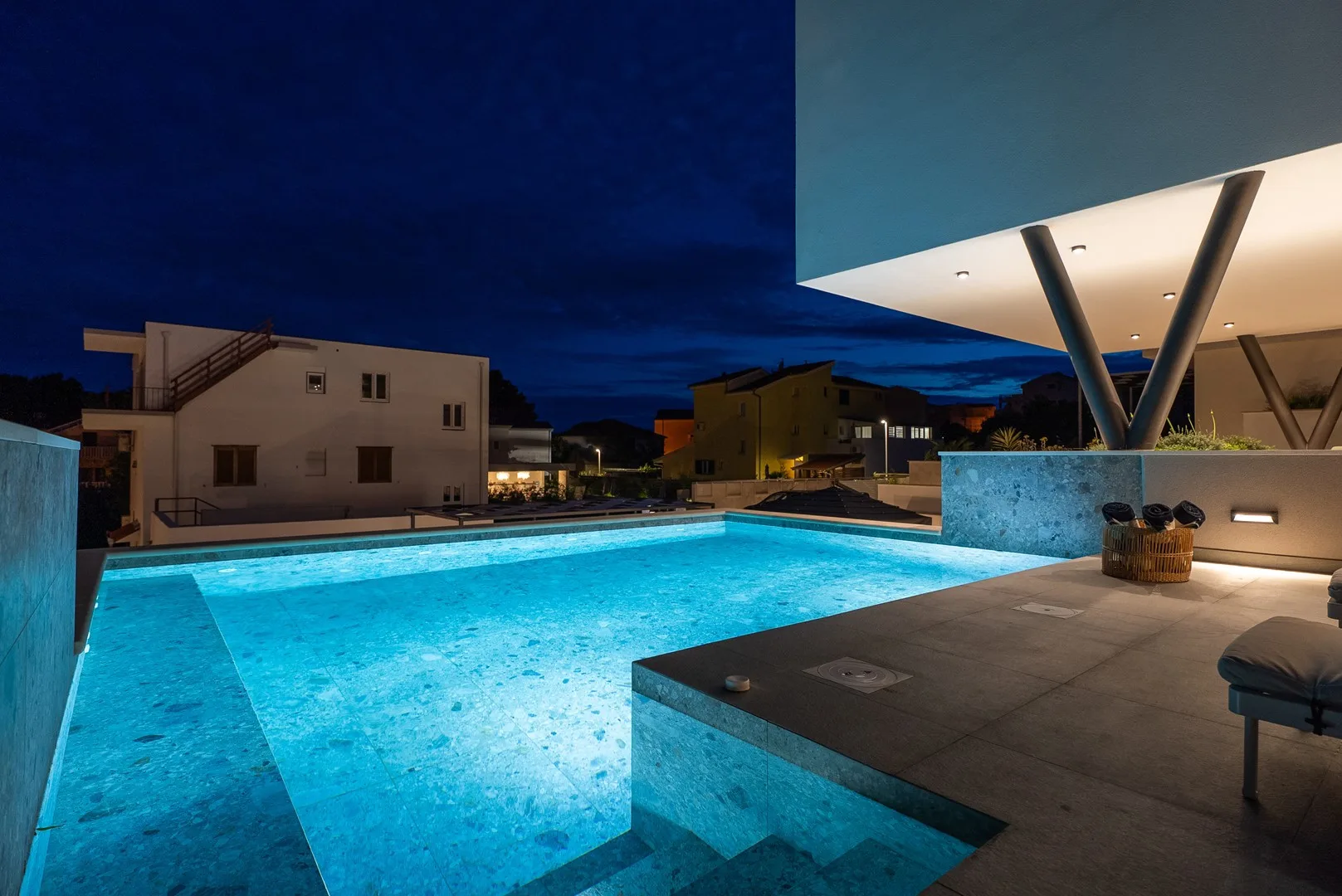 An illuminated outdoor swimming pool beside a modern covered patio at dusk.