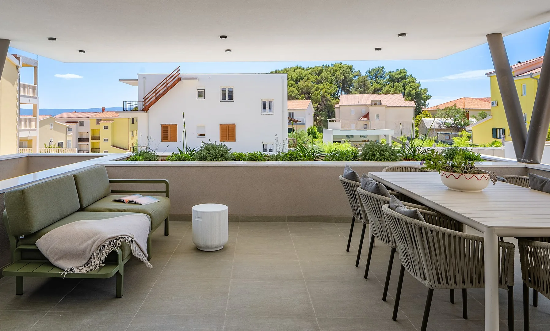 A covered terrace with seating and a dining table overlooks nearby buildings and trees.