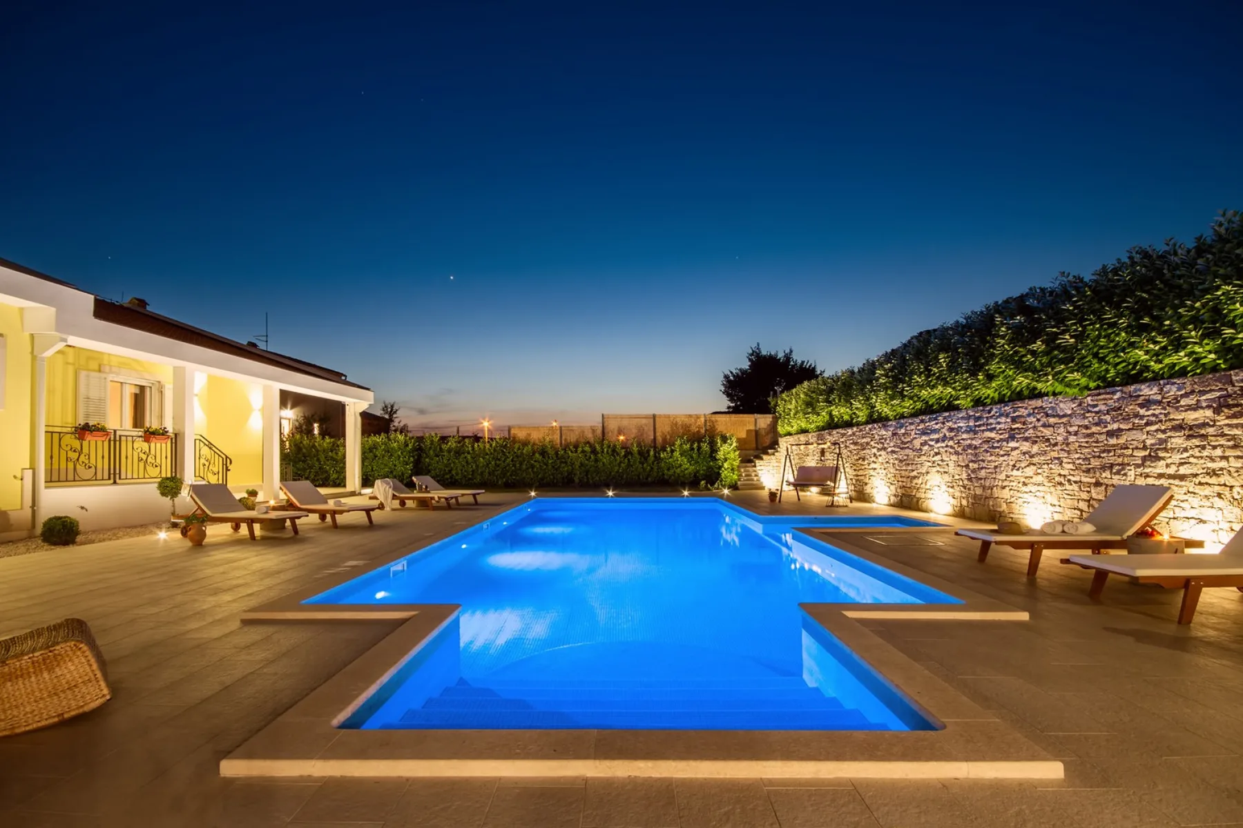 Una piscina al aire libre está rodeada por un patio, asientos y un muro de piedra.