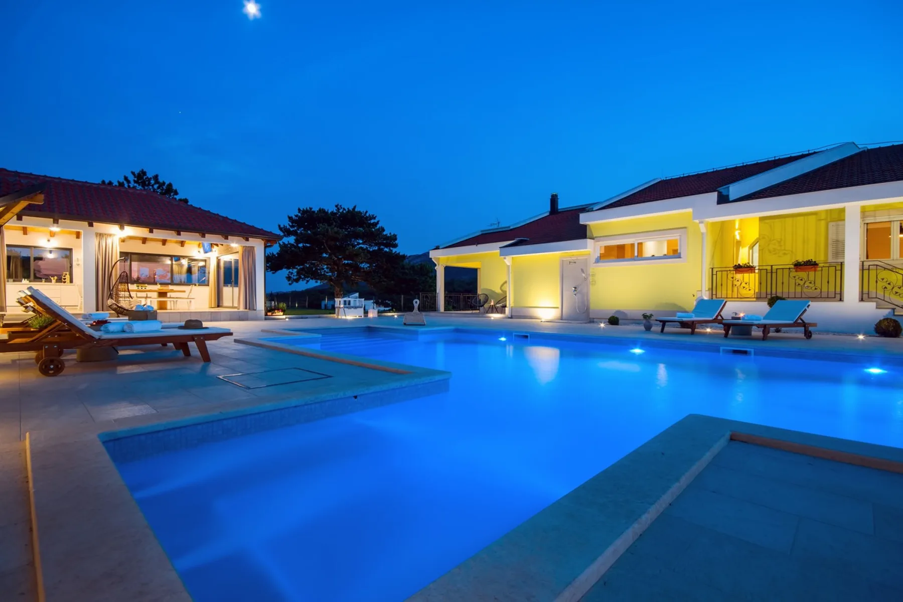 Una piscina brilla en azul al anochecer junto a edificios iluminados.