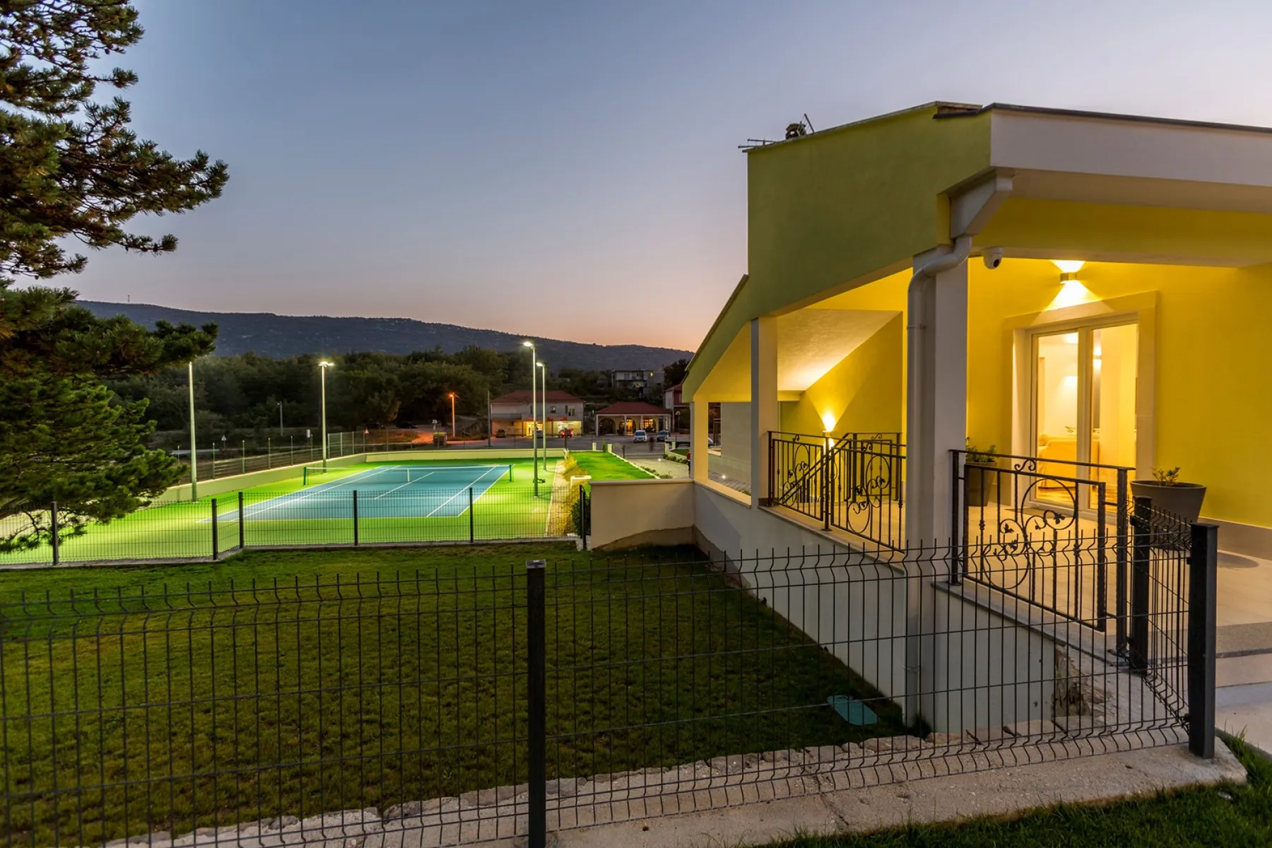 Un edificio iluminado da a una pista de tenis cercada al anochecer.