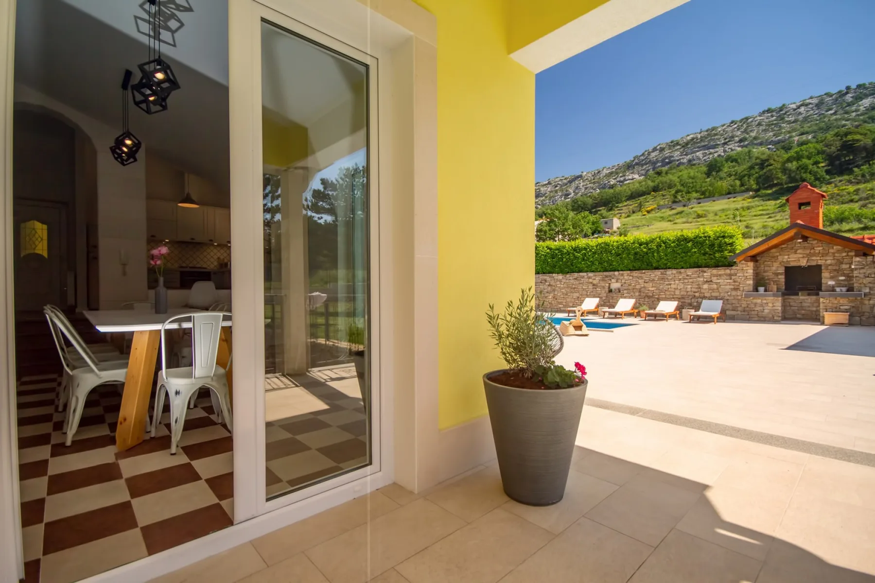 Un patio junto a un comedor da a una terraza soleada con piscina y colina.