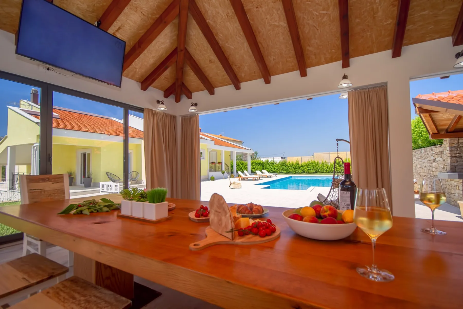 Una mesa de comedor con fruta y bebidas da a una piscina por ventanas abiertas.