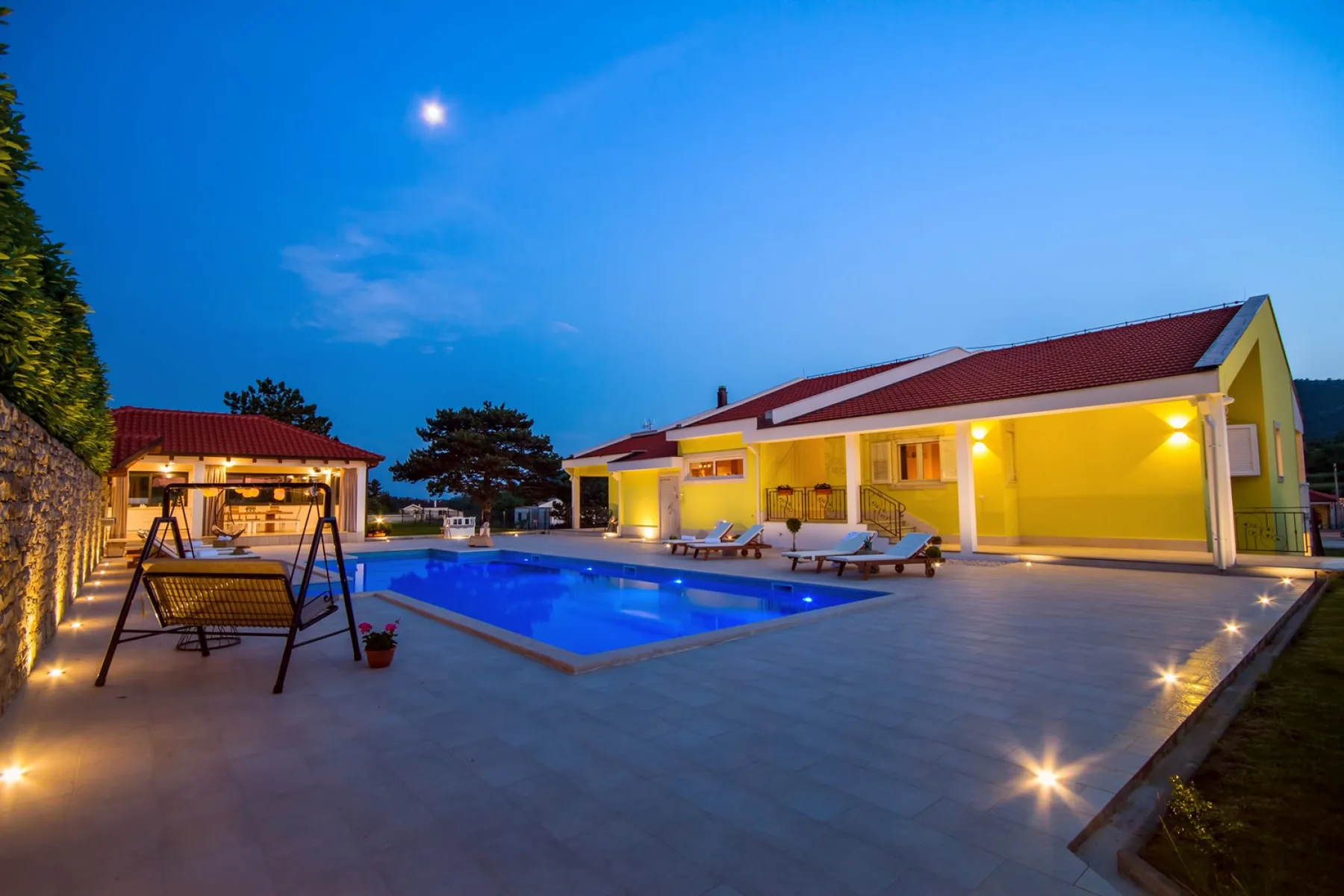 Una piscina iluminada y un patio junto a una casa al anochecer.