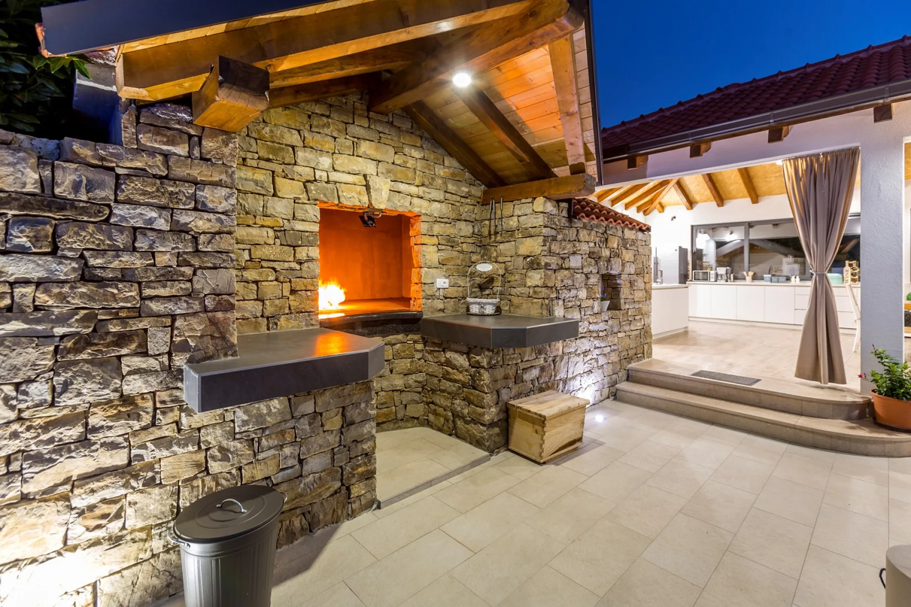 Una zona de cocina exterior de piedra con un horno encendido da a una cocina luminosa.