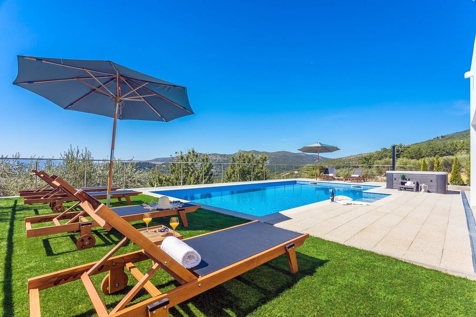 An outdoor swimming pool is surrounded by wooden loungers, umbrellas, and a paved deck.