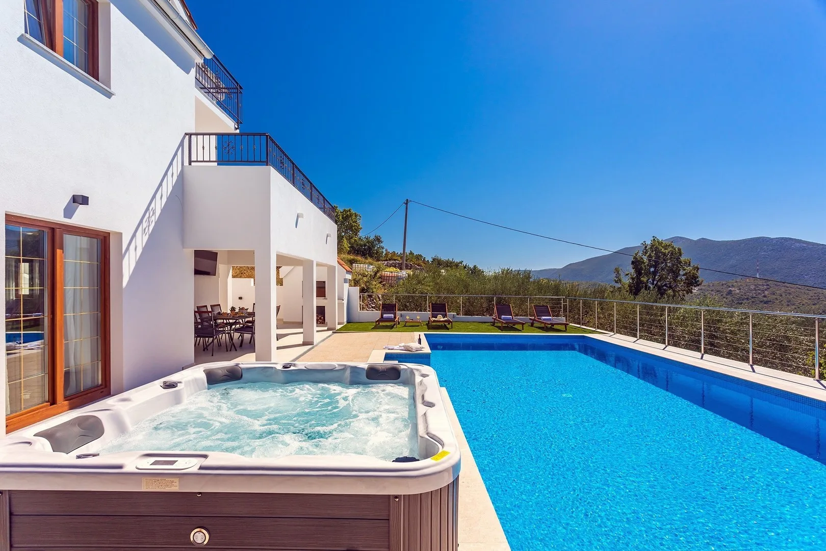An outdoor pool and hot tub beside a white house, with hills in the background.