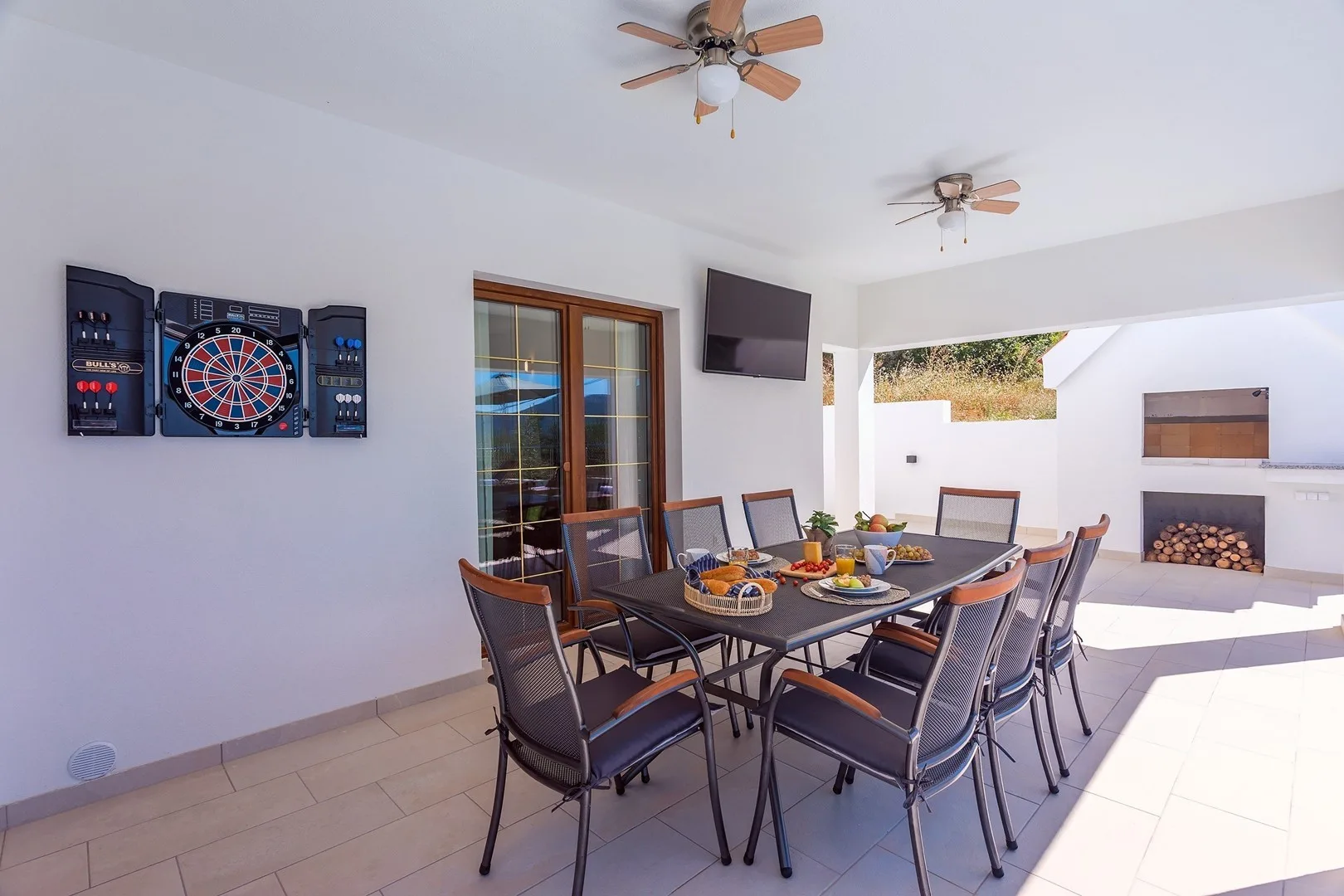 A covered patio dining area has a long table and a dartboard on the wall.