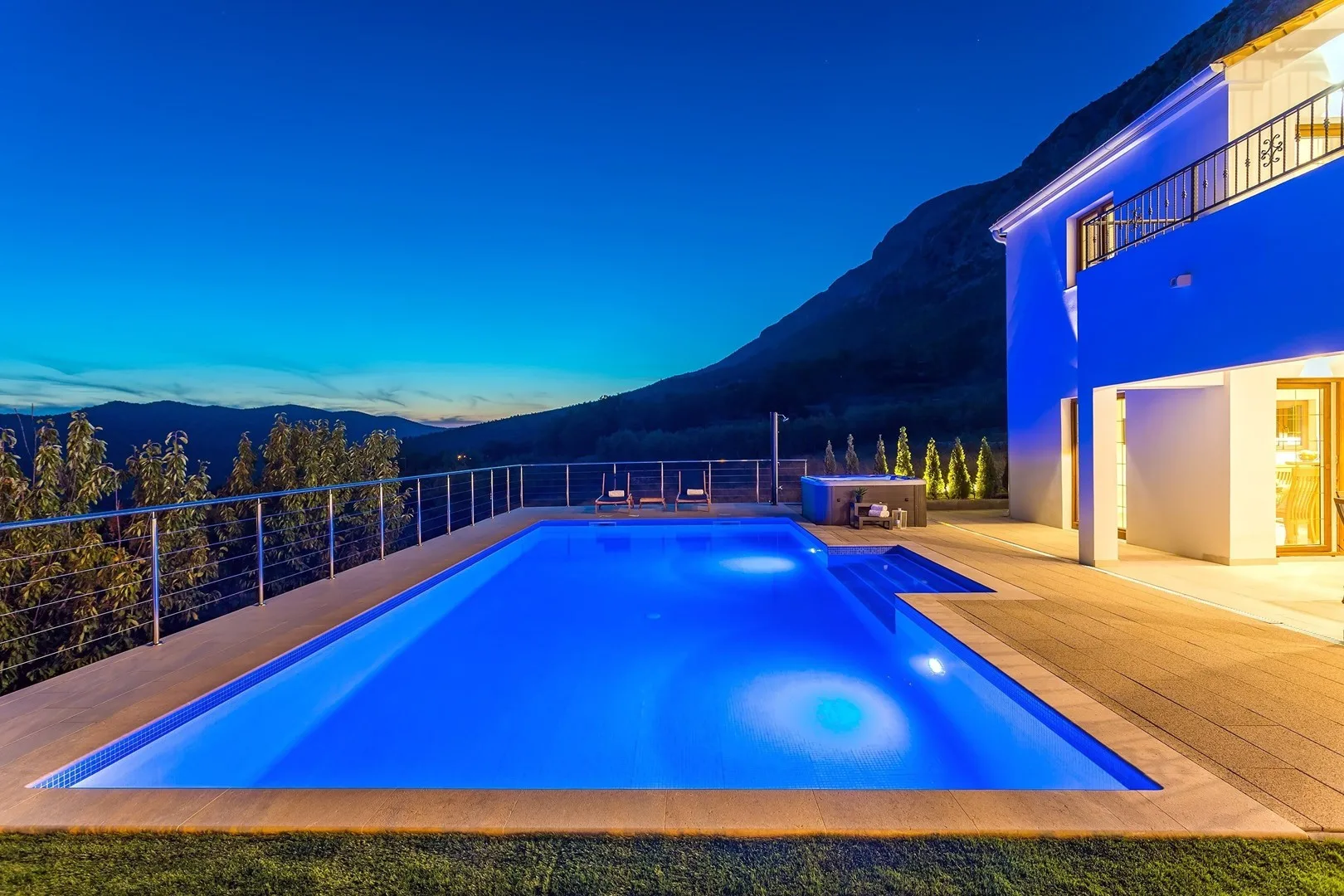 An illuminated swimming pool beside a modern building, with mountain views at dusk.