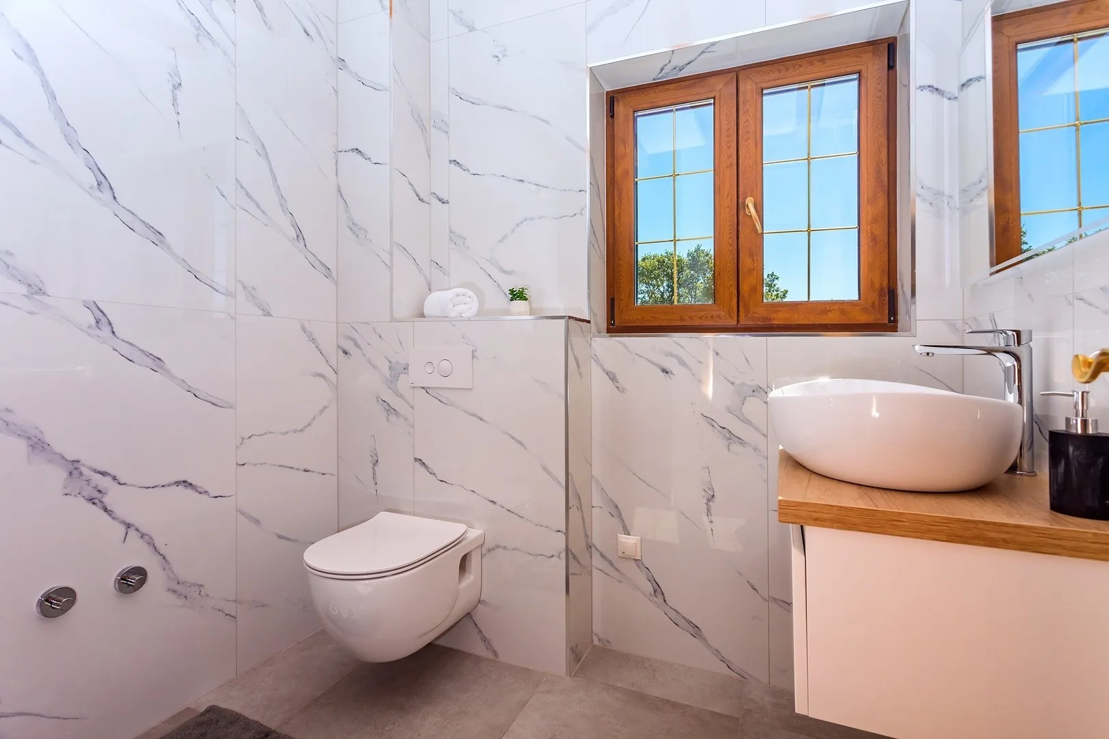 A marble-tiled bathroom with a toilet, sink, and wooden-framed window.