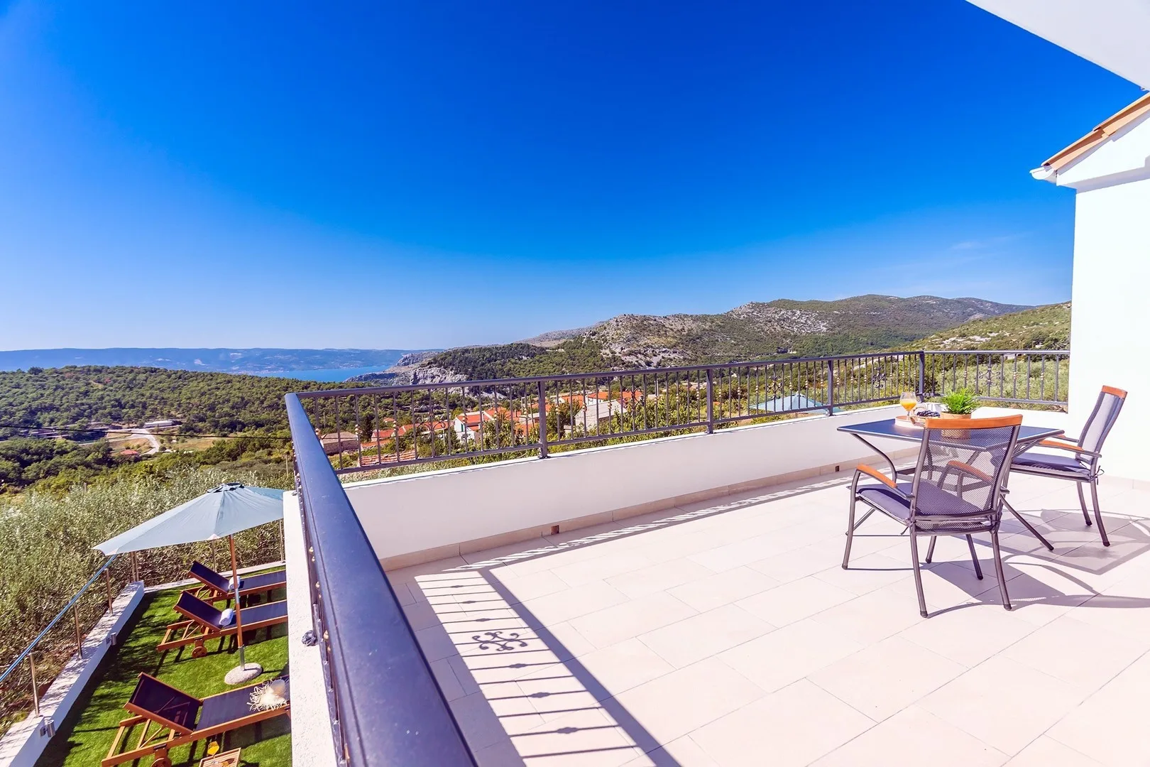 A sunny terrace with outdoor seating overlooks a green hillside and distant water.