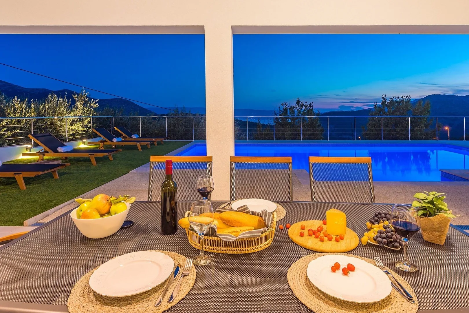 A dining table with plates and food overlooks a swimming pool at dusk.